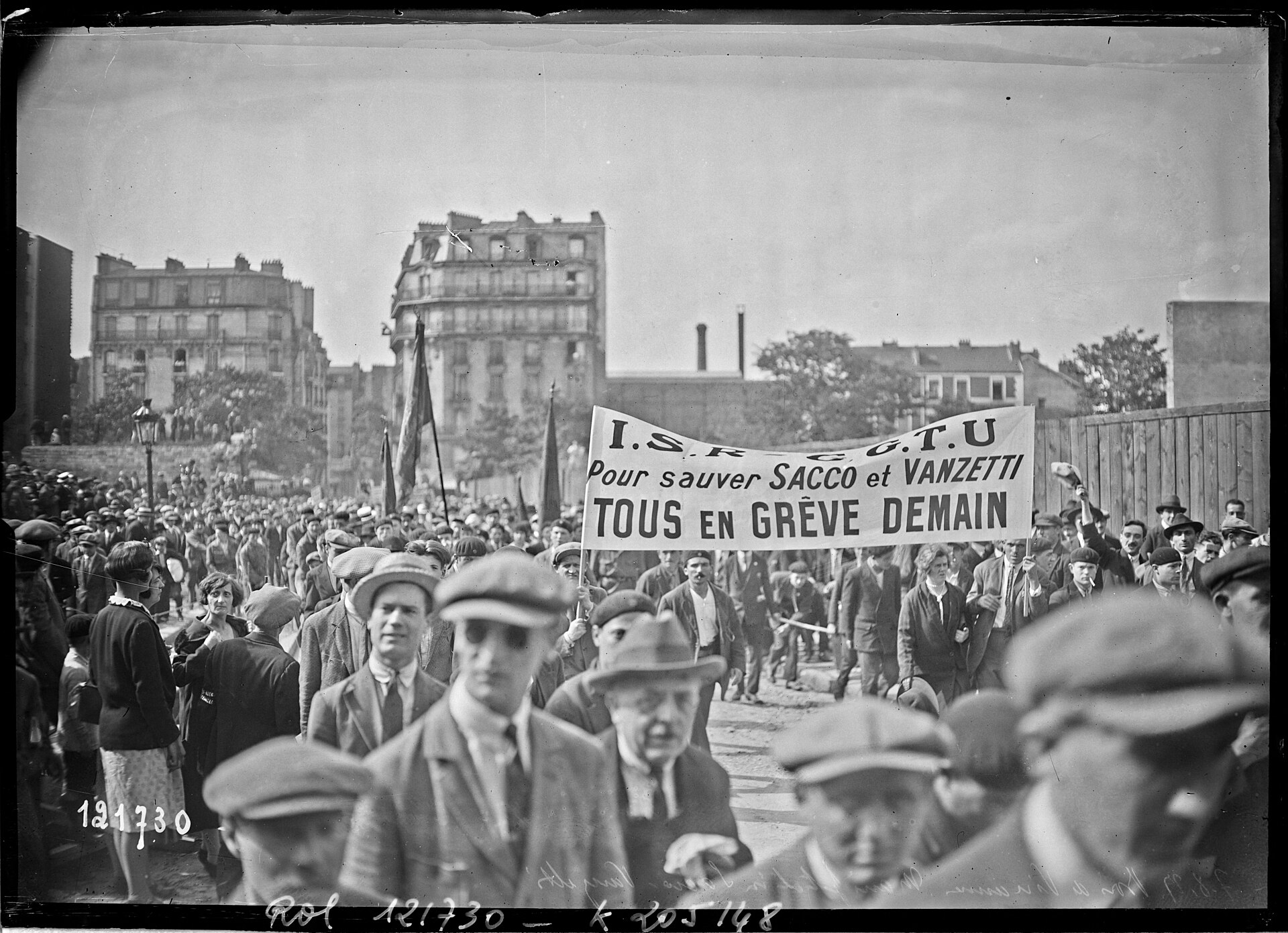 Fotografía en blanco y negro de una multitud de hombres con gorra en una explanada, con una gran pancarta que dice Pour sauver Sacco et Vanzetti, tous en grève demain