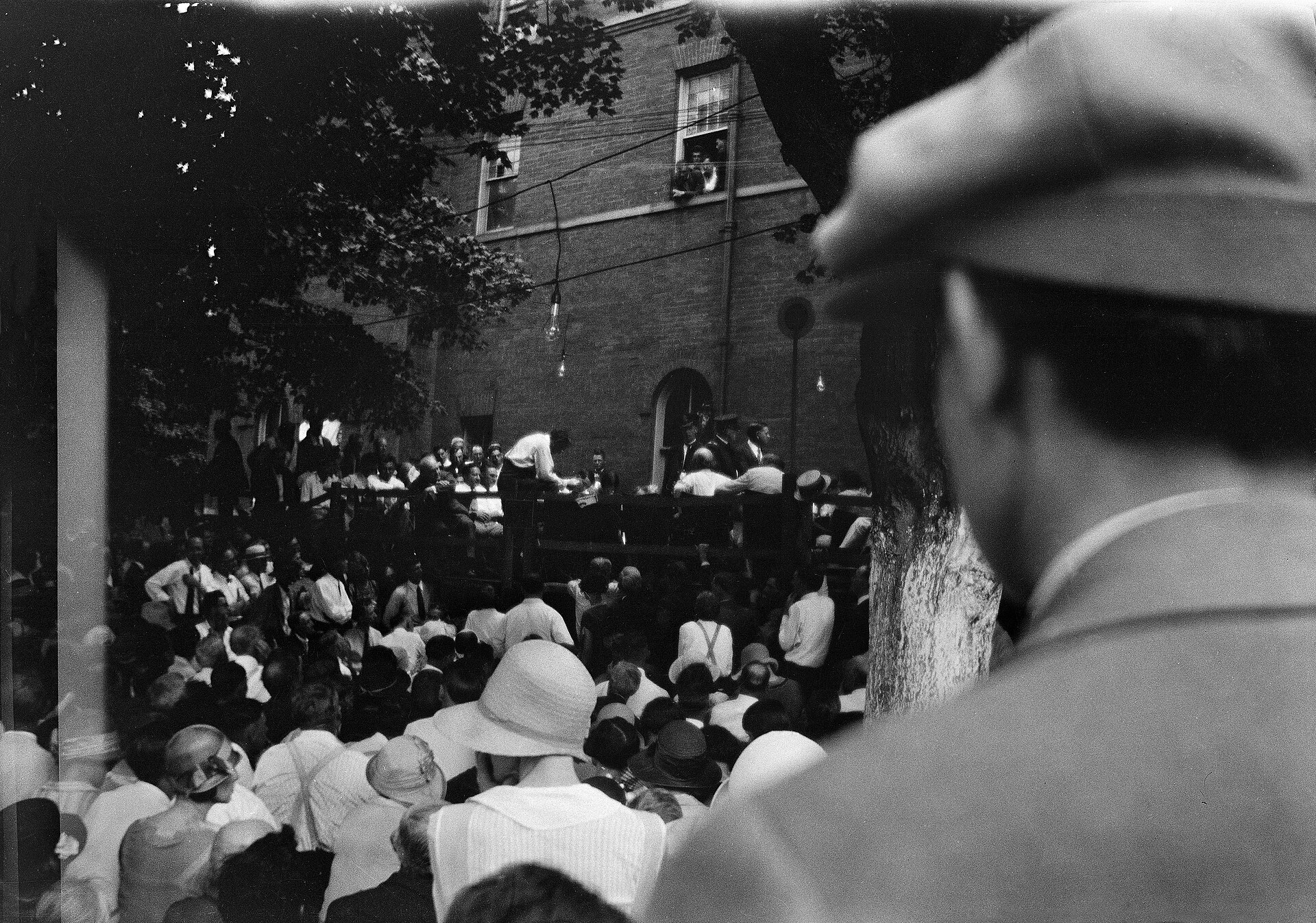Fotografía en blanco y negro tomada al aire libre con una multitud de hombres en mangas de camisa y sombreros de paja rodeando a dos hombres sentados, bajo los árboles