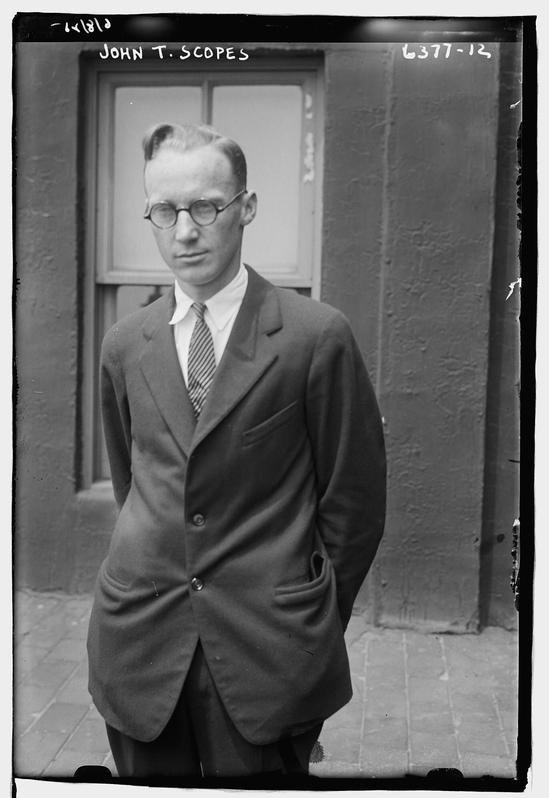 Fotografía en blanco y negro de un hombre joven delgado con gafas redondas, traje y corbata, de pie ante una puerta