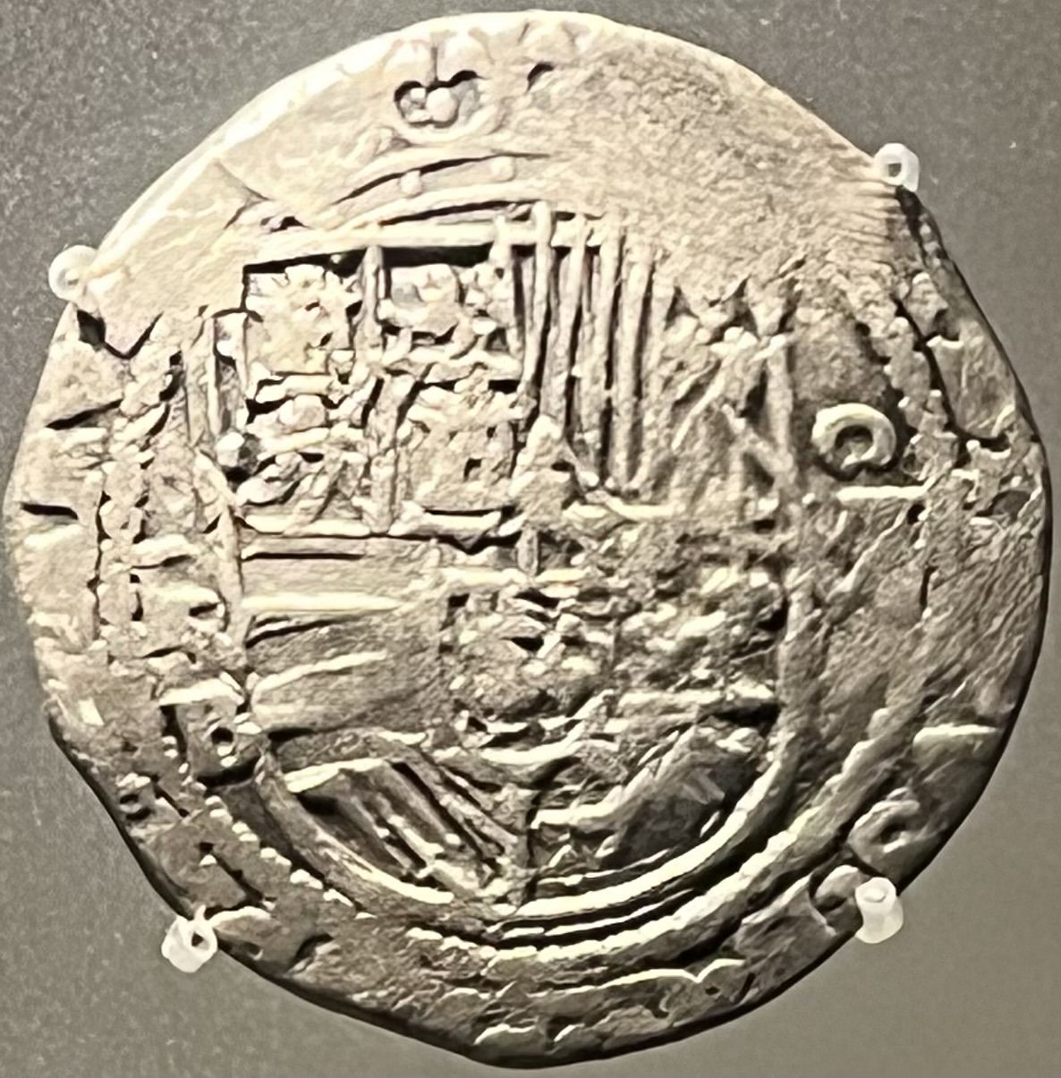 Moneda de plata de forma irregular, acuñada a golpe de martillo, con un escudo cuartelado apenas legible, expuesta en una vitrina