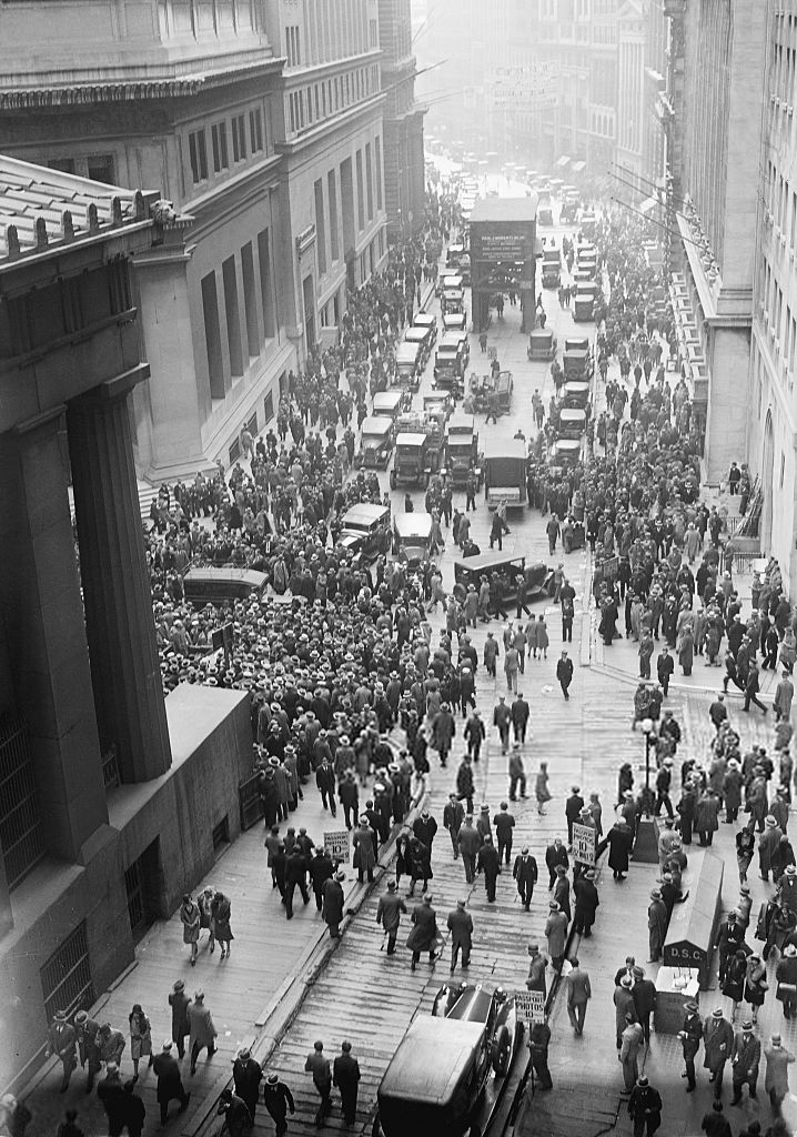 Fotografía en blanco y negro tomada desde lo alto de una calle estrecha de Nueva York llena de gente frente a un edificio con columnas, con coches detenidos en el centro de la calzada