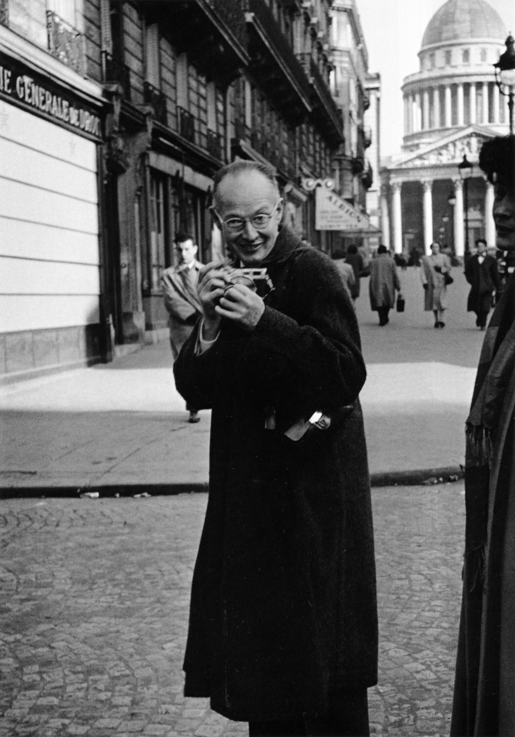 Henri Cartier-Bresson, con gafas y abrigo oscuro, sonríe con una pequeña cámara Leica entre las manos en una calle de París; al fondo se ve la cúpula del Panteón