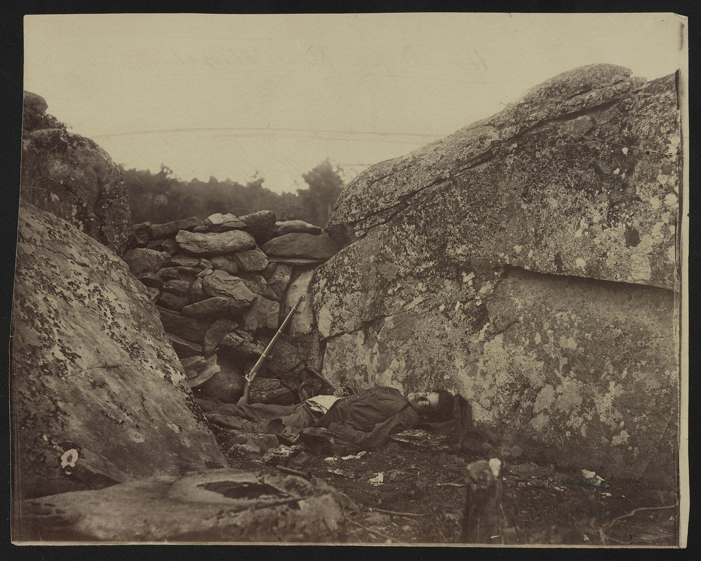 Fotografía de 1863 del cadáver de un soldado tendido entre grandes rocas, con un fusil apoyado contra el muro de piedra