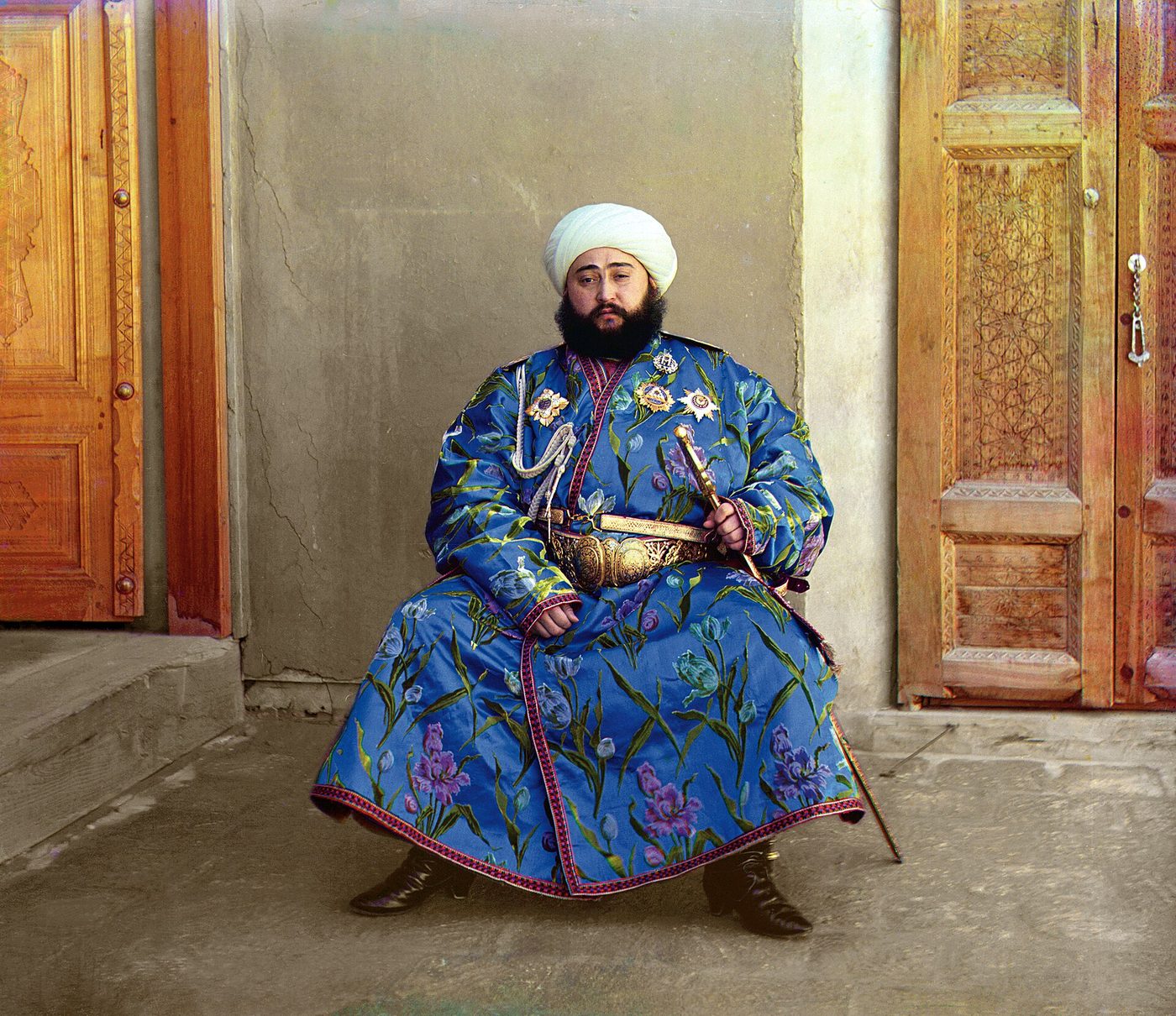 Fotografía en color intenso de un hombre con turbante blanco y túnica azul bordada, sentado con la mano en la empuñadura de un sable