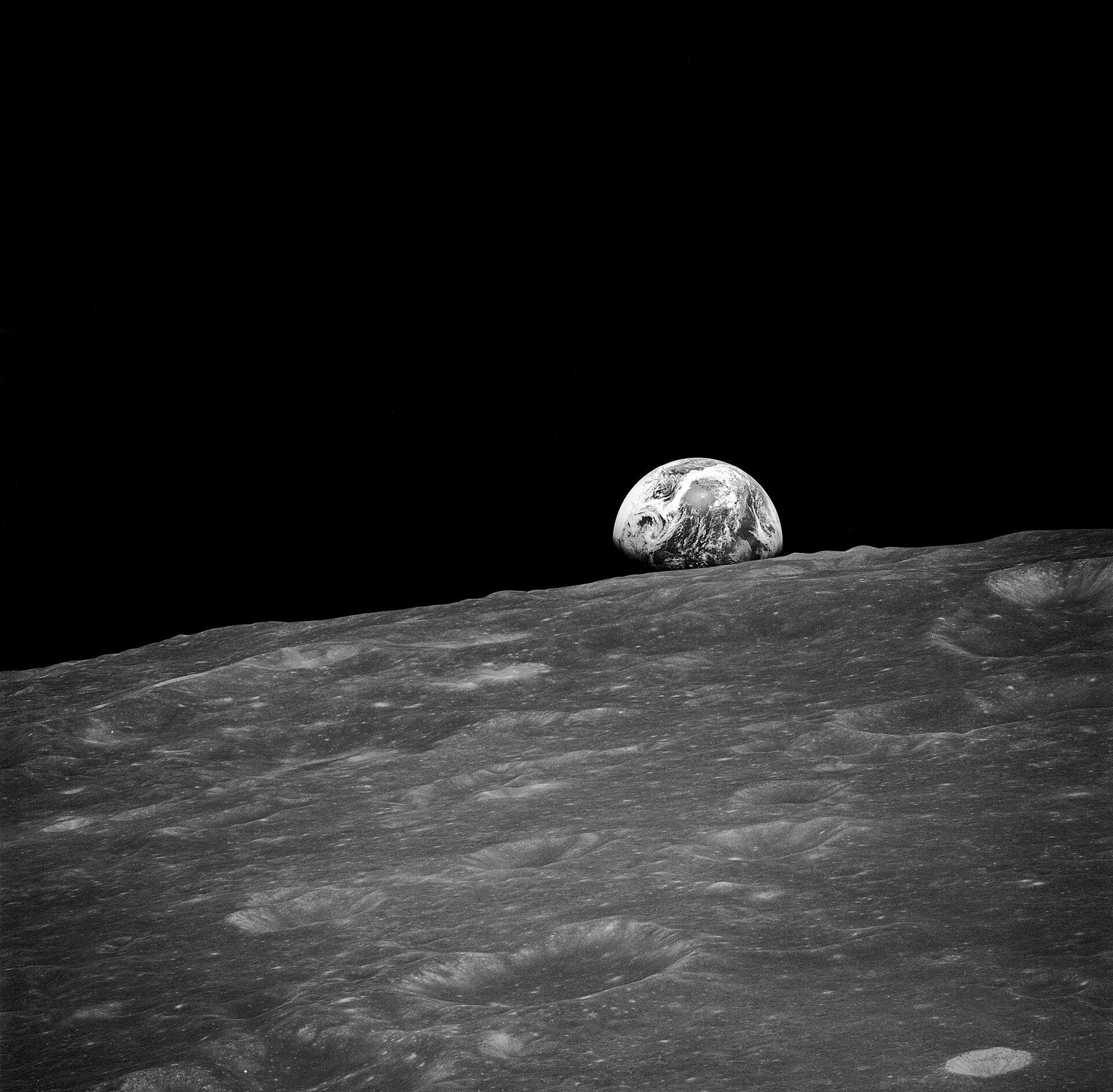 Fotografía en blanco y negro del horizonte lunar con cráteres en primer plano y, al fondo, sobre el cielo negro, una Tierra parcialmente iluminada