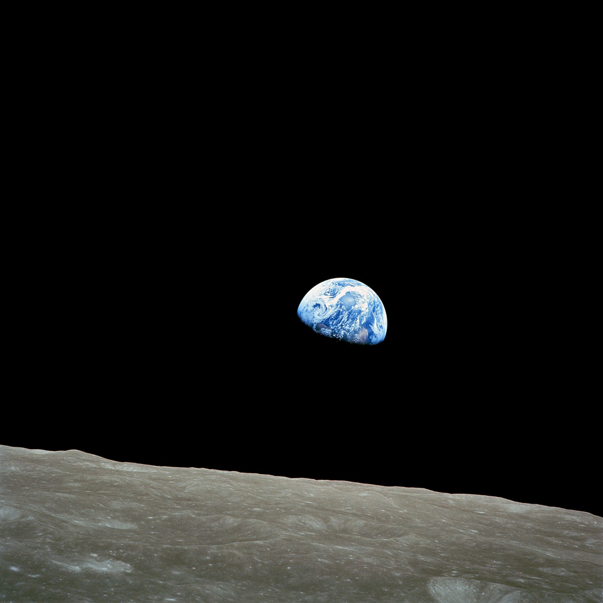 Fotografía en color de la Tierra, media esfera azul con nubes blancas, sobre el negro del espacio, por encima del horizonte gris y lleno de cráteres de la Luna
