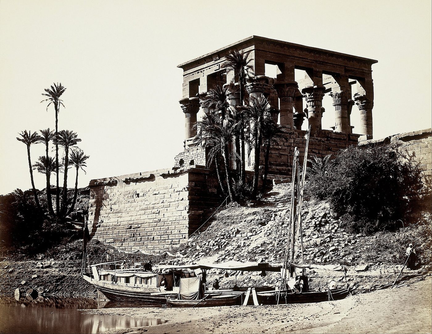 Fotografía de hacia 1857 de un templo egipcio con columnas sobre la orilla del Nilo, con palmeras y una barca amarrada