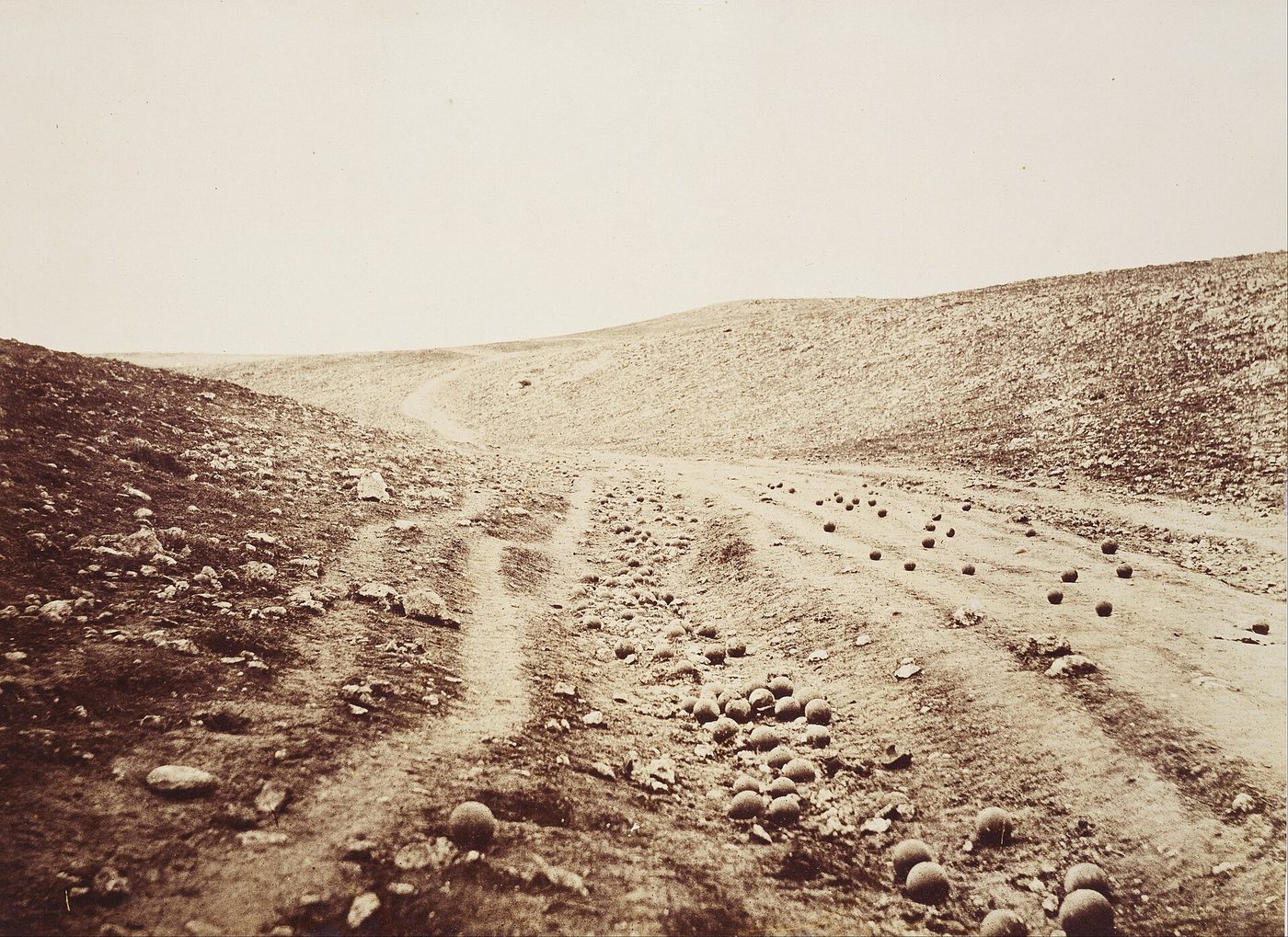 Fotografía de 1855 de un camino de tierra que atraviesa una hondonada árida entre dos lomas, con decenas de balas de cañón esféricas esparcidas por la calzada