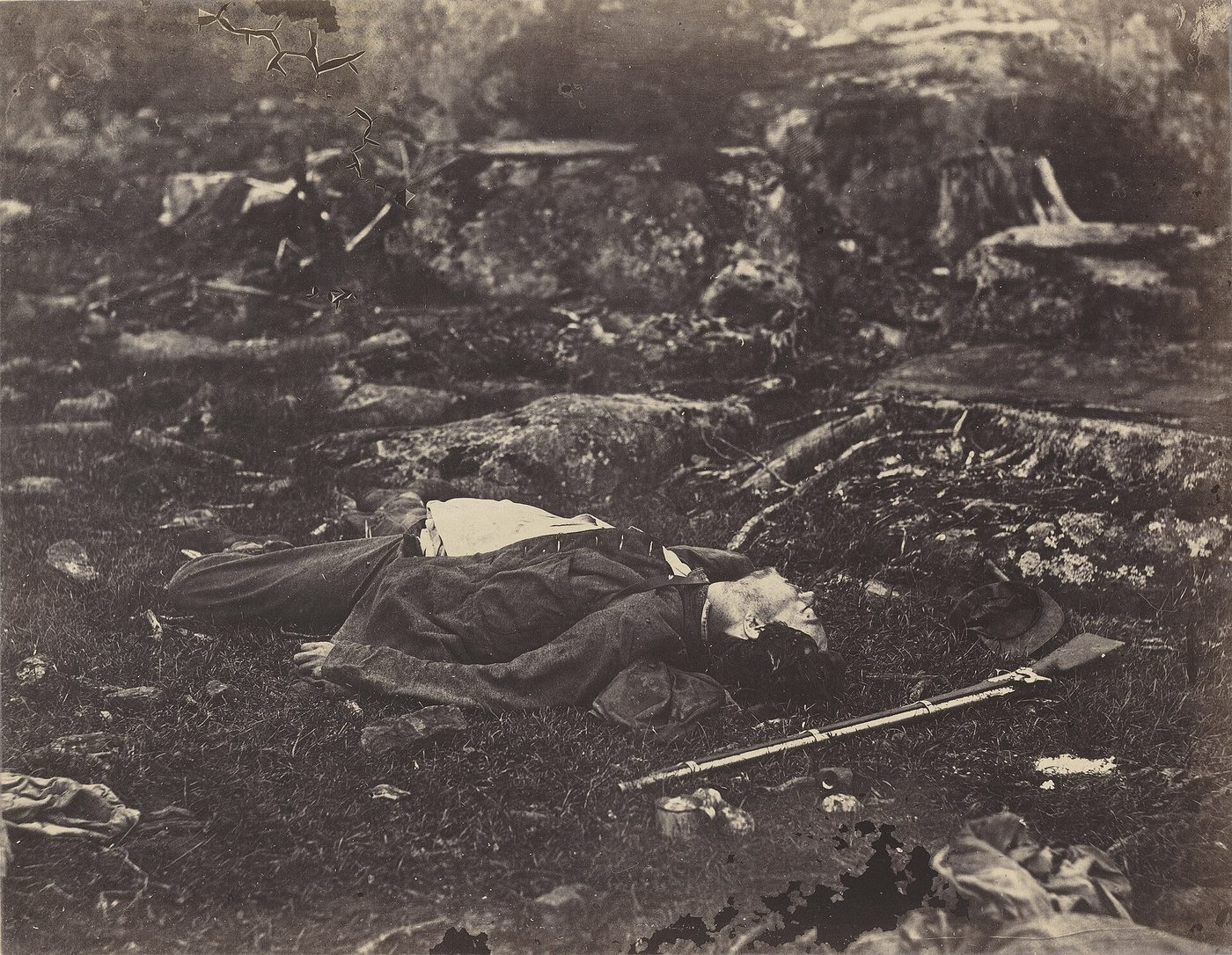 Fotografía de 1863 de un soldado muerto tendido en una ladera de hierba y piedras, con la cara vuelta hacia el cielo y un fusil a sus pies