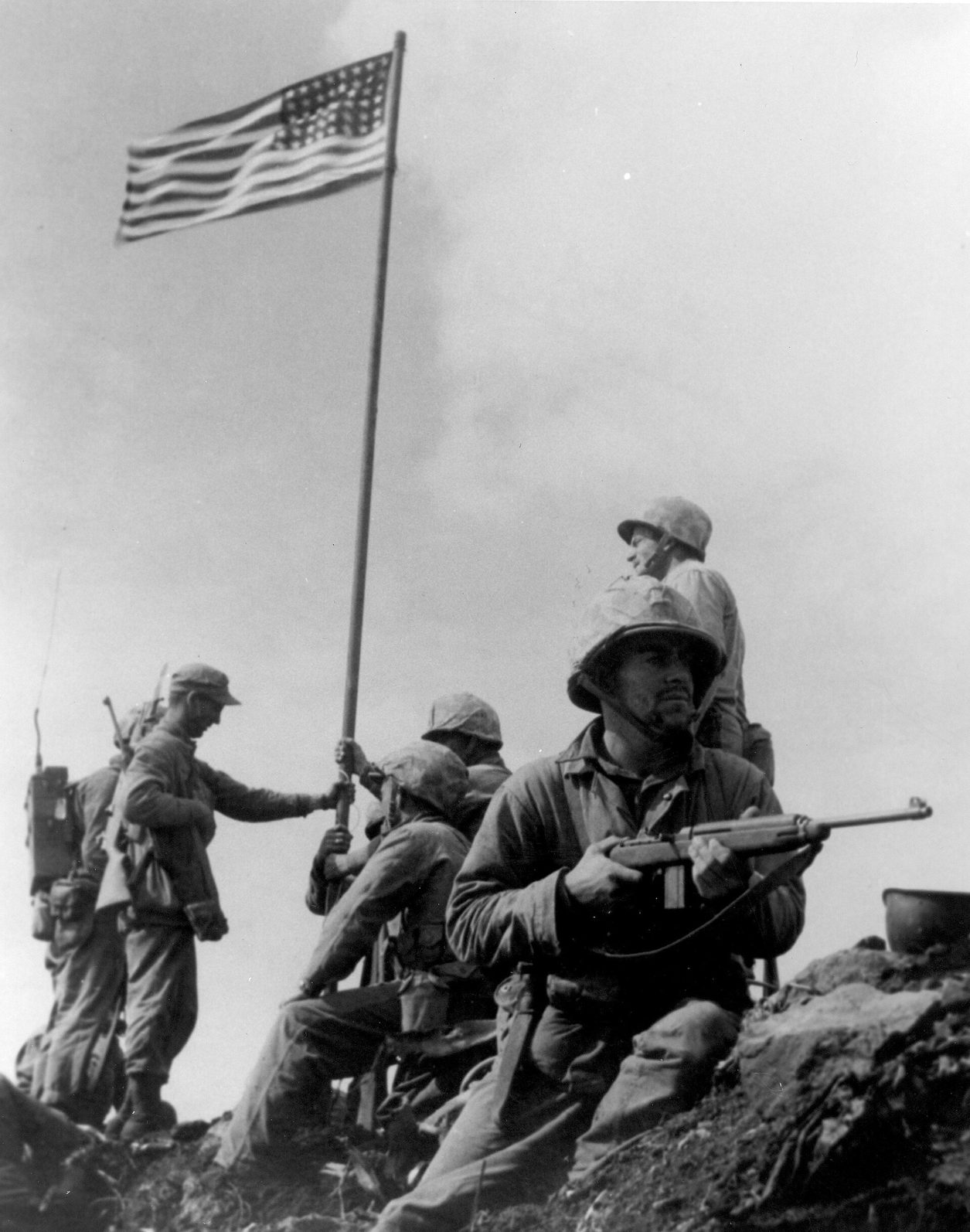 Fotografía en blanco y negro de un grupo de marines en la cima de una colina; varios sujetan un mástil con una bandera pequeña de Estados Unidos y uno, en primer plano, empuña una carabina