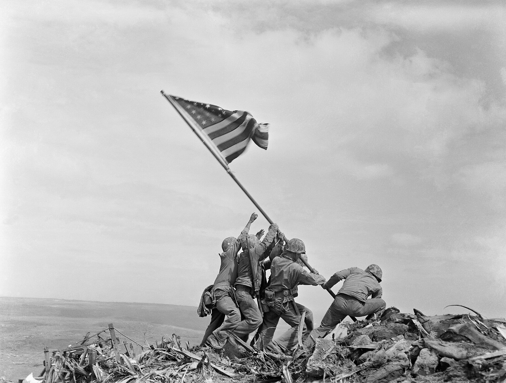 Fotografía en blanco y negro de seis soldados que levantan entre todos un mástil largo con una bandera de Estados Unidos ondeando, sobre un terreno de escombros, con el cielo nublado detrás