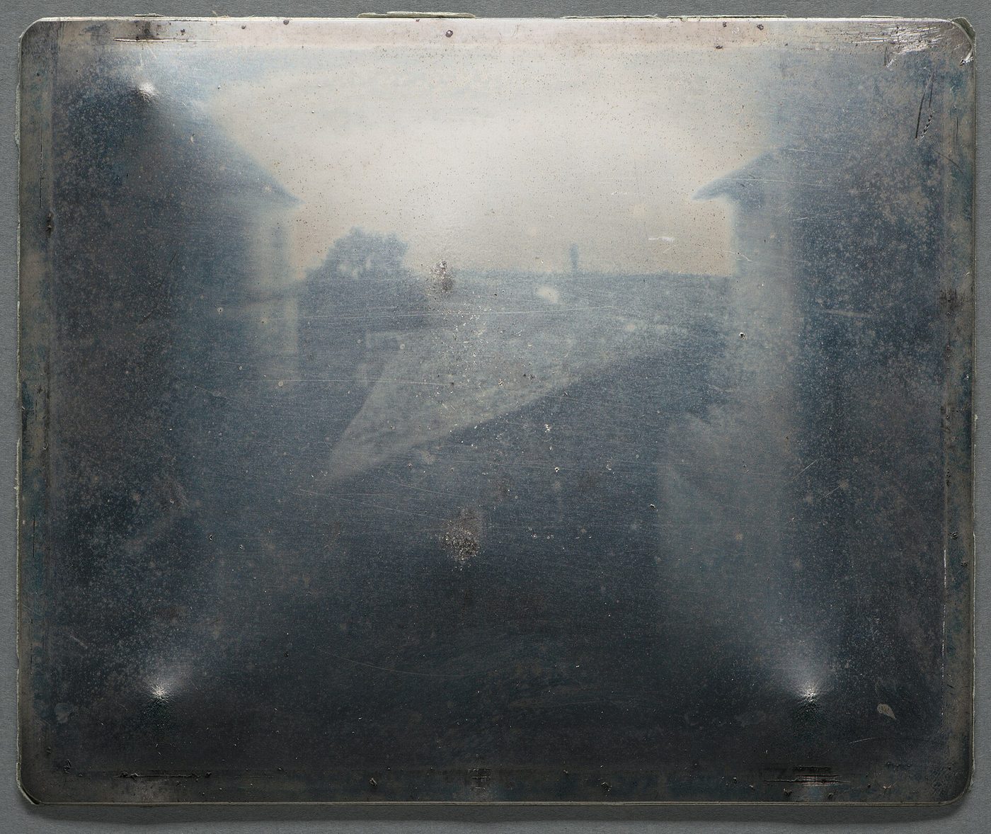 Placa metálica gris azulada, rectangular, con reflejos de luz en las esquinas y una imagen muy tenue de tejados y edificios apenas distinguible