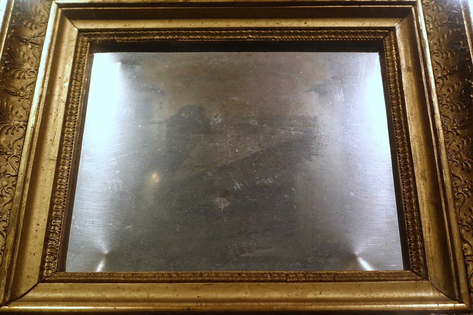 Marco dorado antiguo que contiene una placa metálica brillante, casi espejada, dentro de una vitrina de museo