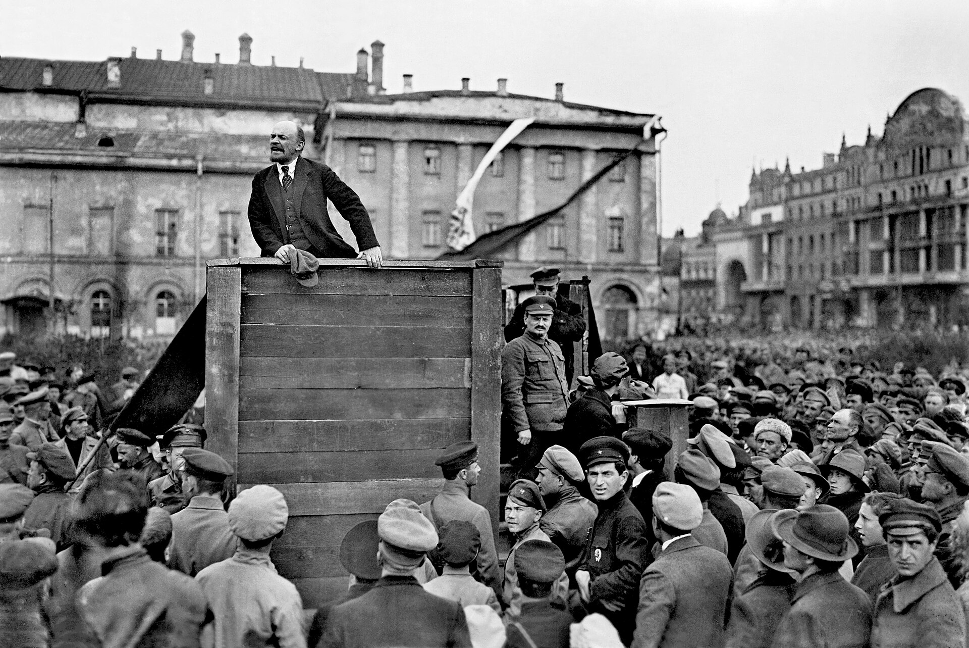 Fotografía de 1920 de Lenin hablando desde una tribuna de madera ante una multitud de soldados con gorras, frente a un gran edificio; en los escalones de la derecha hay varios hombres de pie