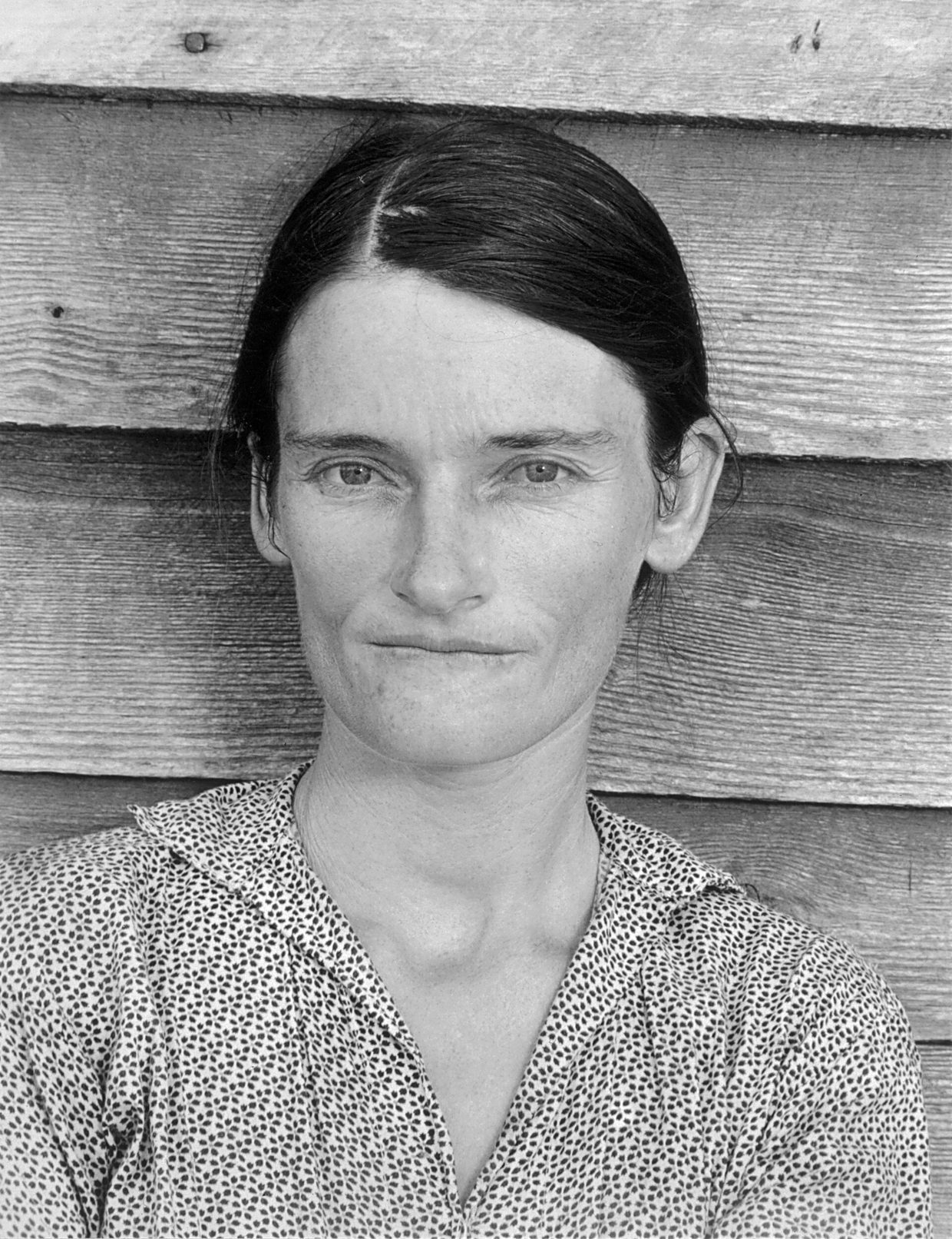 Retrato frontal de 1936 de una mujer joven con vestido de cuadros de pie ante una pared de tablas de madera, mirando directamente a cámara
