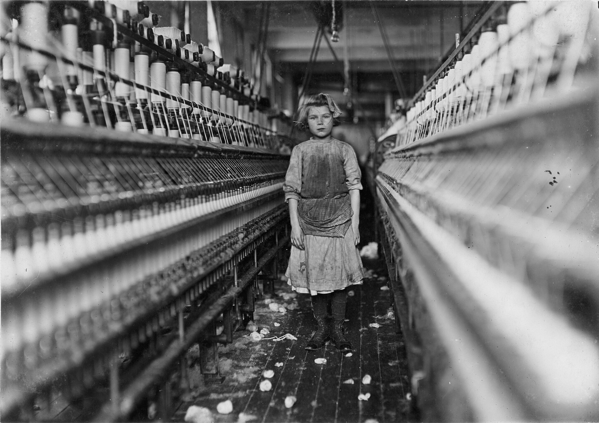Fotografía de 1909 de una niña descalza de pie junto a una larga hilera de husos en una fábrica textil