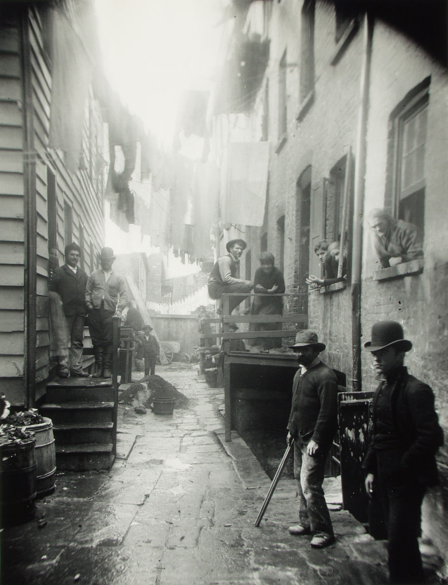 Fotografía de 1888 de un callejón estrecho de Nueva York con ropa tendida en lo alto y varios hombres apoyados en las paredes mirando a cámara