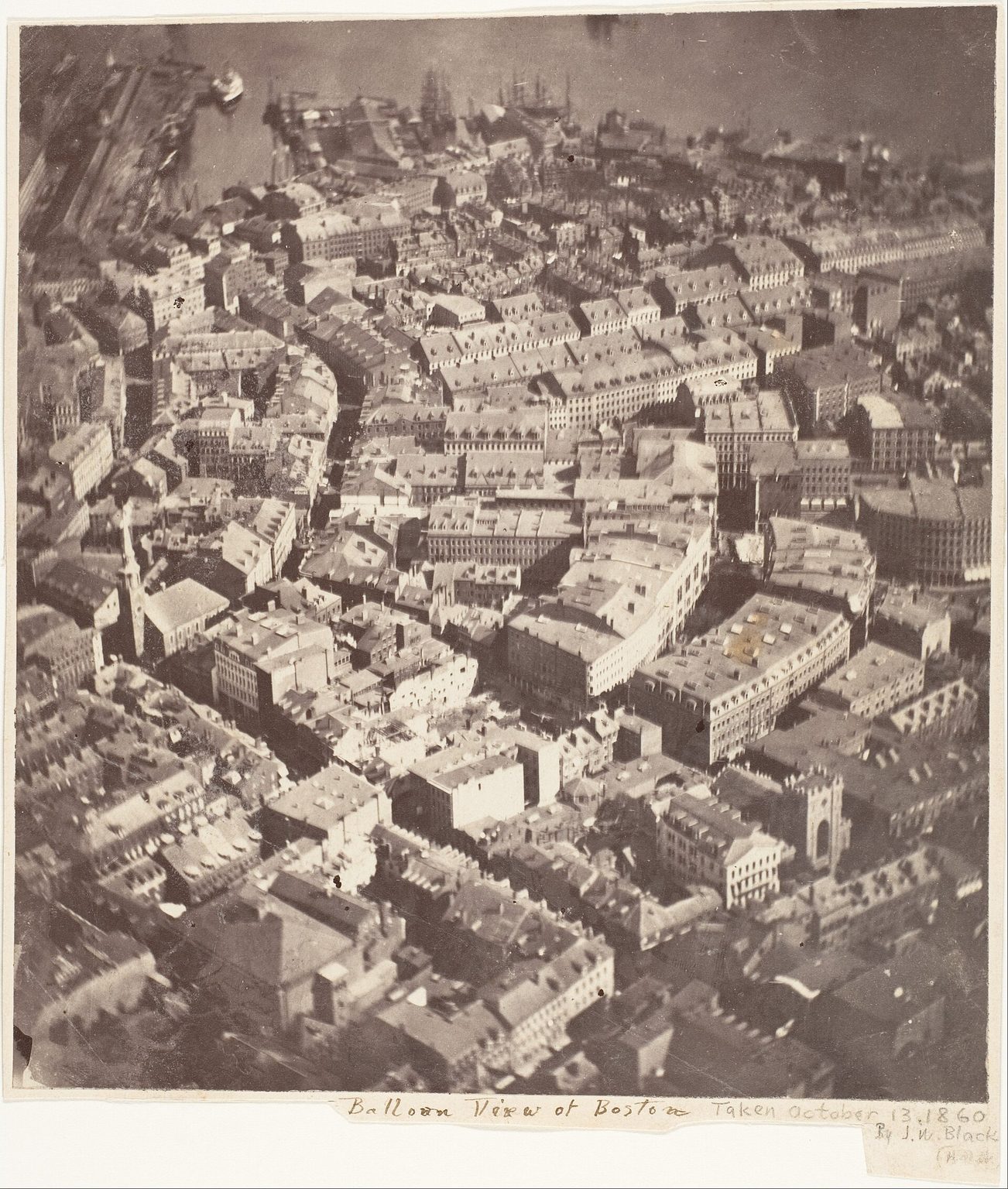 Fotografía antigua en tonos sepia de una ciudad vista desde lo alto, con tejados, calles estrechas, torres de iglesias y muelles al fondo