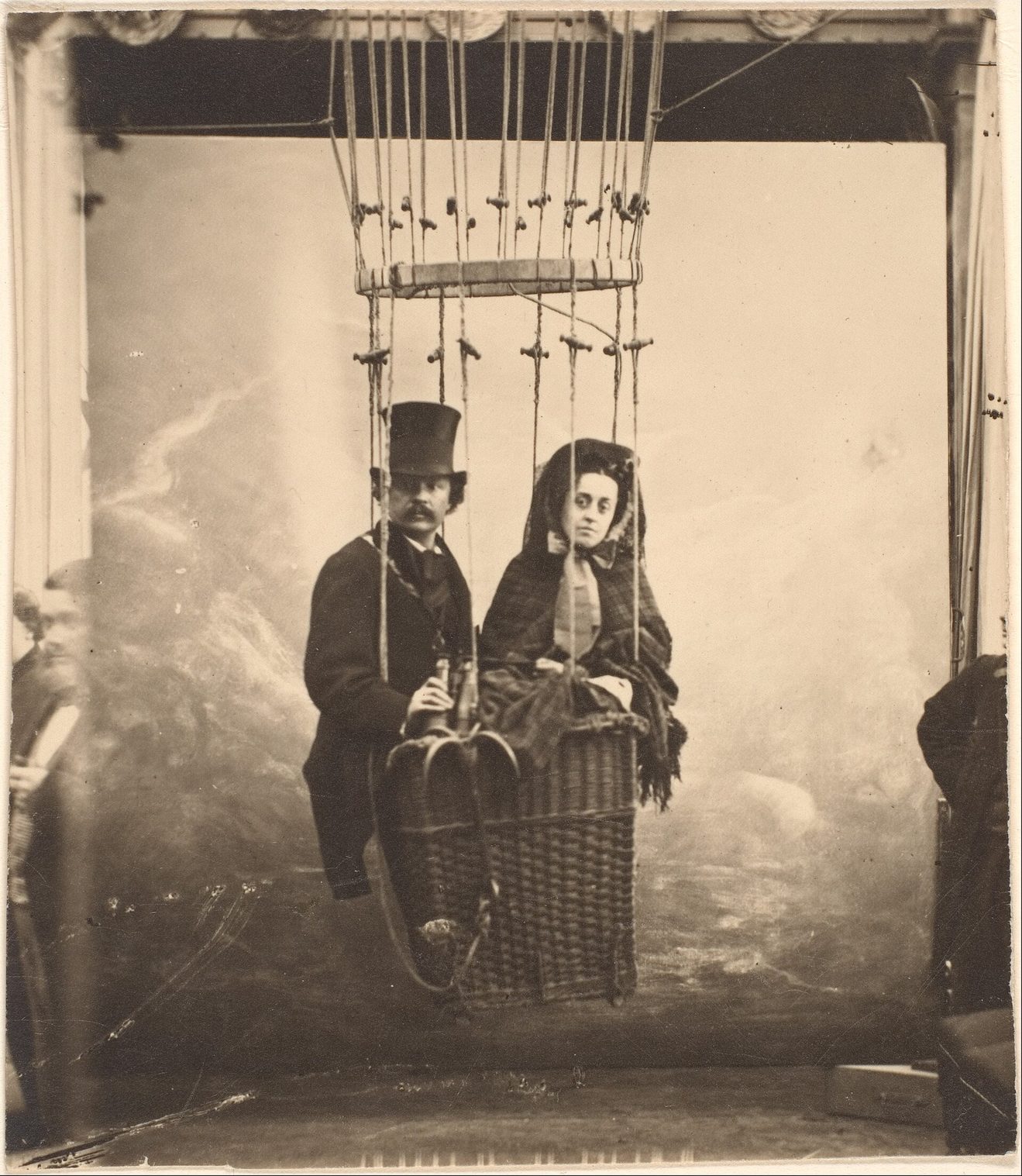 Fotografía de estudio de un hombre con sombrero de copa y una mujer vestida de negro sentados en la barquilla de un globo, colgada de cuerdas ante un fondo pintado de nubes