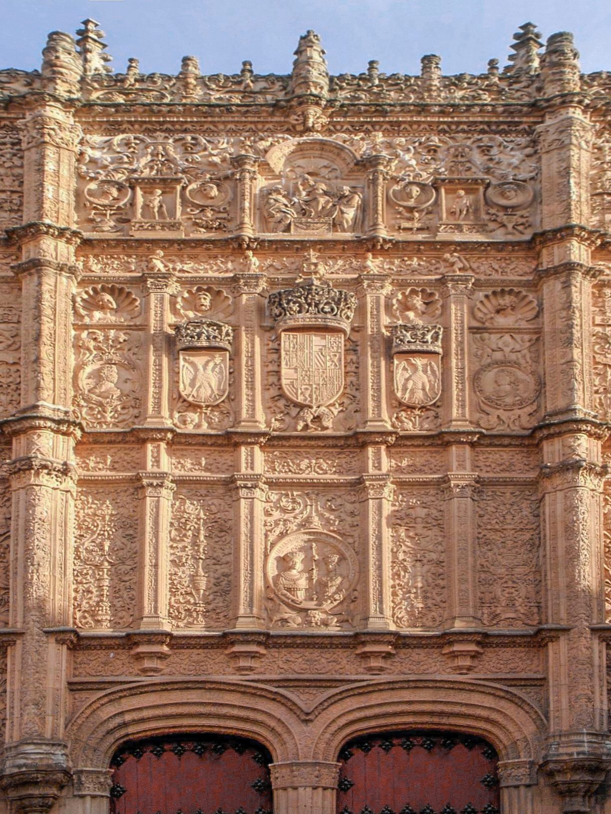 Fachada plateresca de piedra dorada profusamente labrada, con escudos y medallones, sobre dos puertas en arco