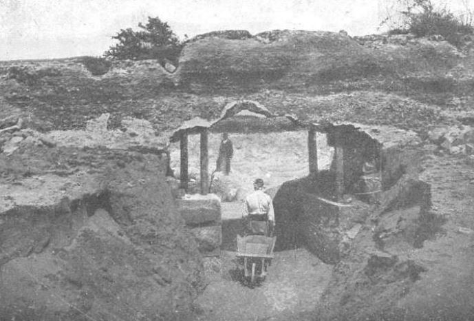 Fotografía antigua en blanco y negro de una zanja de excavación entre muros de tierra y piedra, con un obrero que empuja una carretilla y otro hombre de pie al fondo
