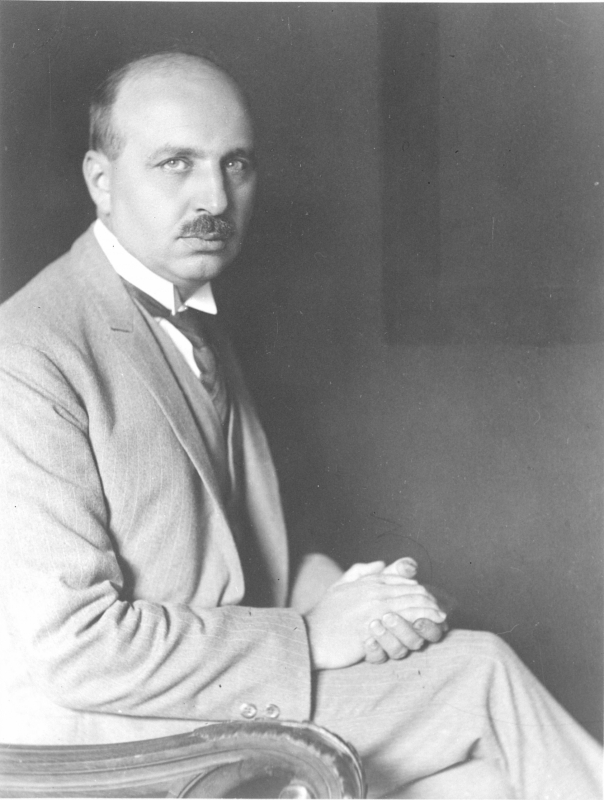 Fotografía en blanco y negro de un hombre calvo con bigote, traje claro y corbata, sentado con las manos cruzadas sobre las rodillas