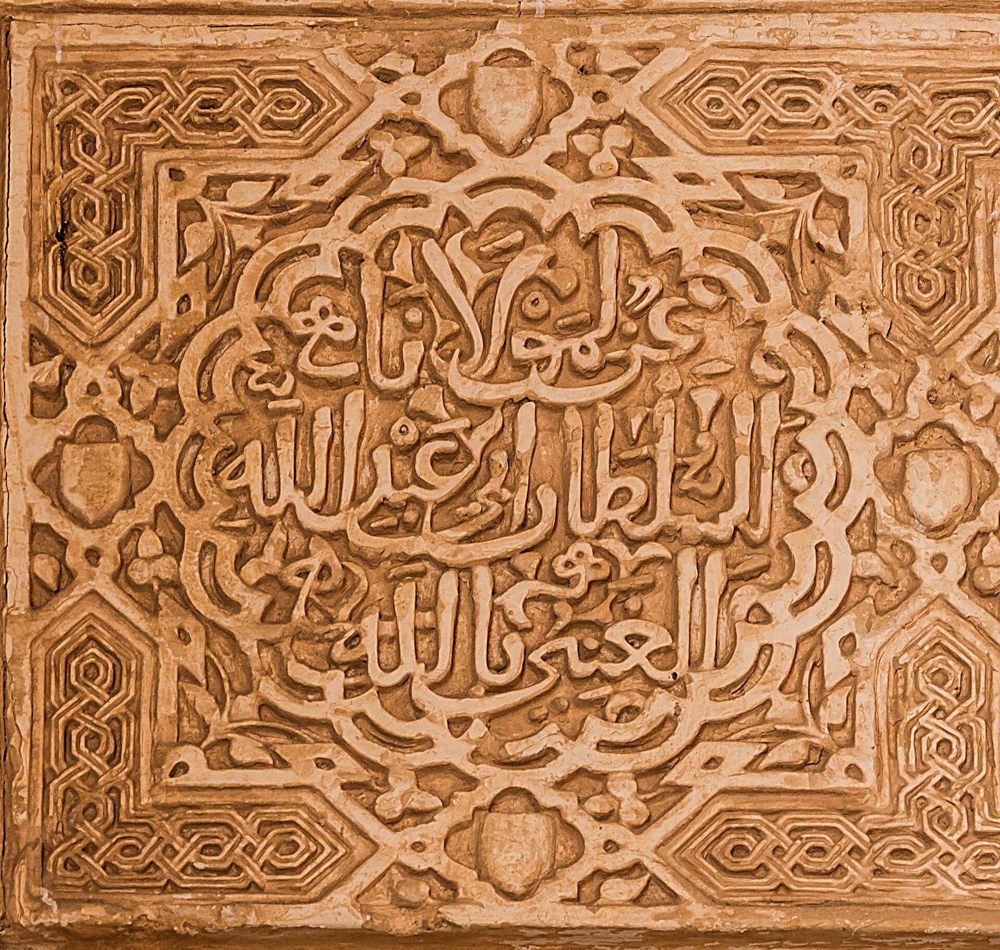 Relieve de yesería color ocre con una inscripción en caligrafía árabe cursiva en el centro, rodeada de un fondo de lacerías y motivos vegetales geométricos