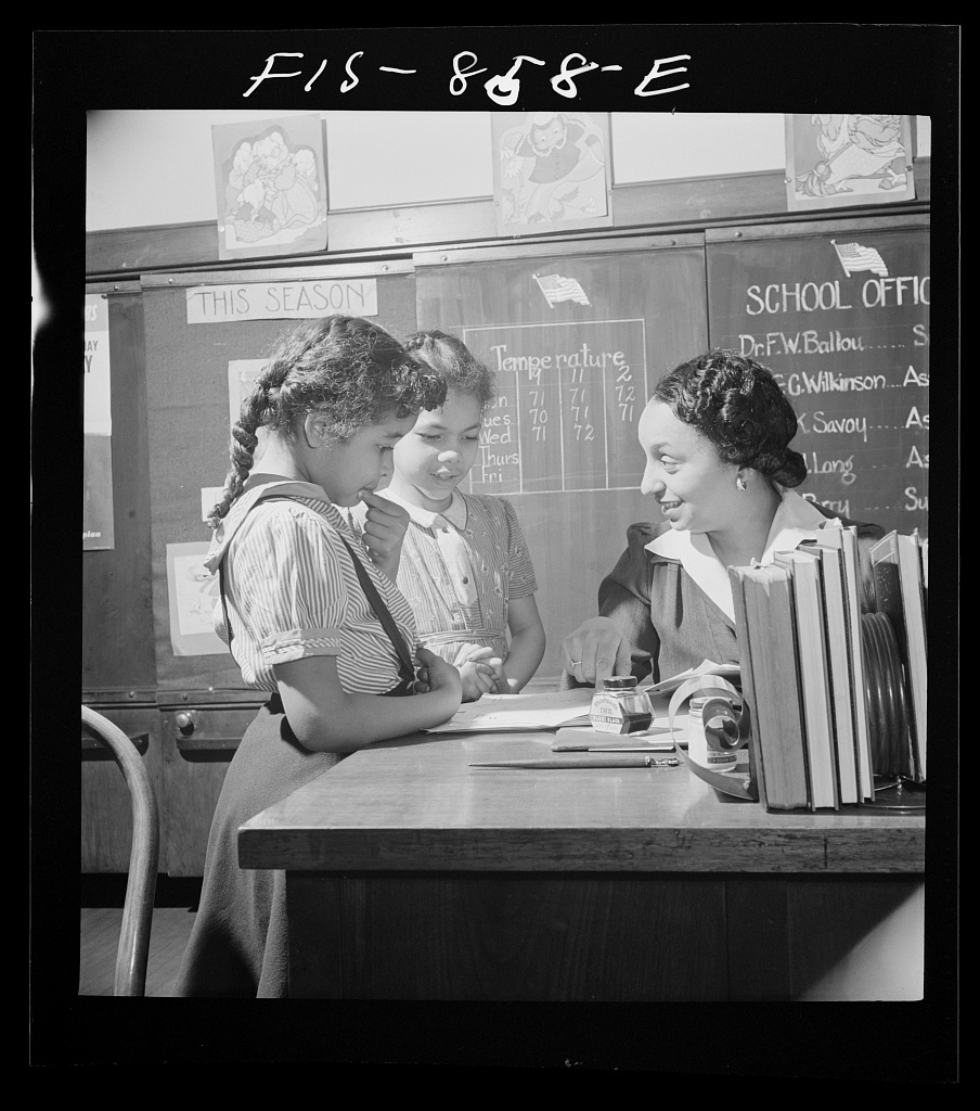Fotografía en blanco y negro de una maestra sentada a una mesa junto a dos niñas de pie que miran un cuaderno, ante una pizarra con carteles
