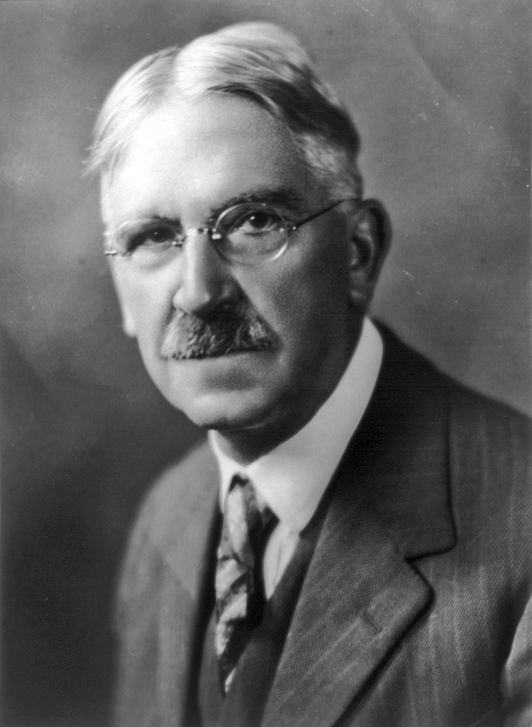 Retrato en blanco y negro de un hombre mayor con gafas redondas, bigote y pelo blanco, con traje, chaleco y corbata