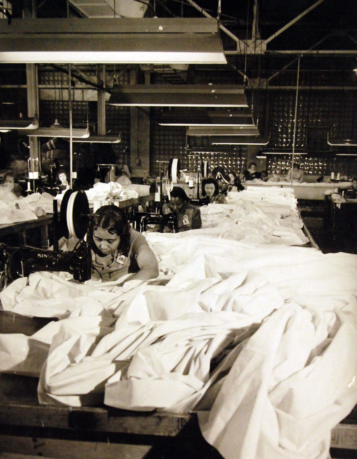 Fotografía en blanco y negro de una nave industrial con largas mesas cubiertas de tela blanca y mujeres cosiendo con máquinas eléctricas