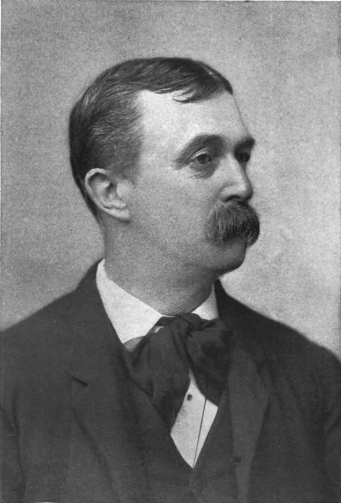 Retrato fotográfico en blanco y negro de un hombre con bigote, traje oscuro y pajarita, de perfil