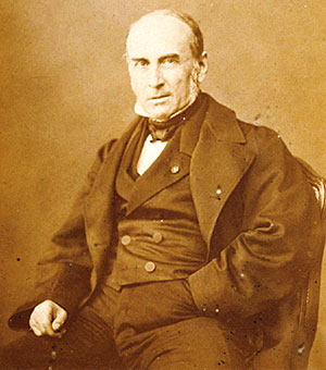 Retrato fotográfico en tono sepia de un hombre mayor calvo, sentado, con levita oscura y corbata de lazo
