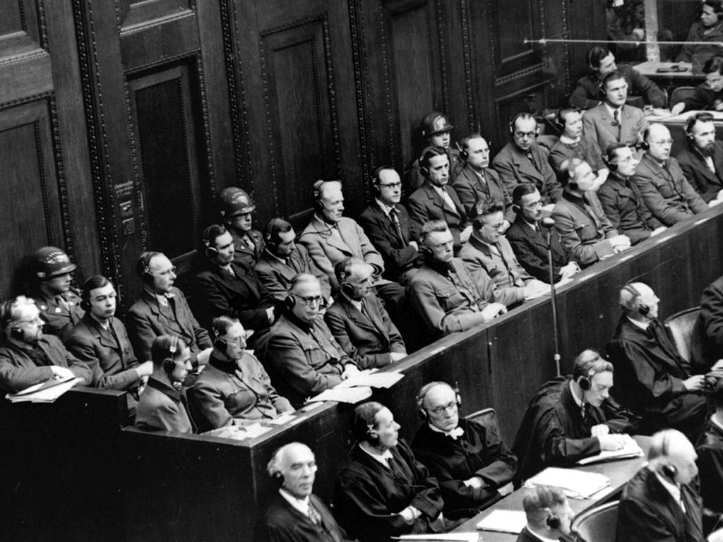 Fotografía en blanco y negro de una sala de tribunal con dos filas de acusados en el banquillo custodiados por soldados, y sus abogados delante