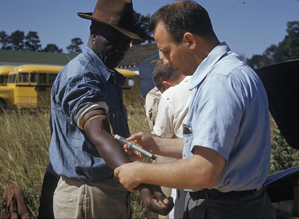 Fotografía en color de un sanitario que extrae sangre del brazo de un hombre negro con sombrero en un campo
