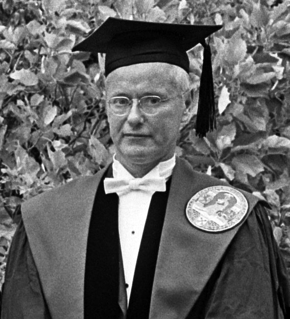 Retrato en blanco y negro de un hombre con gafas, birrete y toga académica con una medalla, ante un fondo de hojas