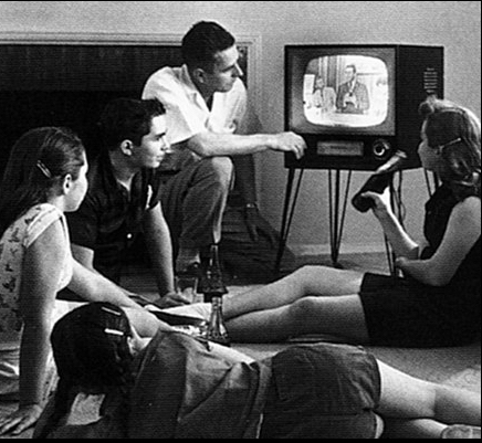 Fotografía en blanco y negro de varias personas sentadas y tumbadas en el suelo de un salón mirando un televisor antiguo