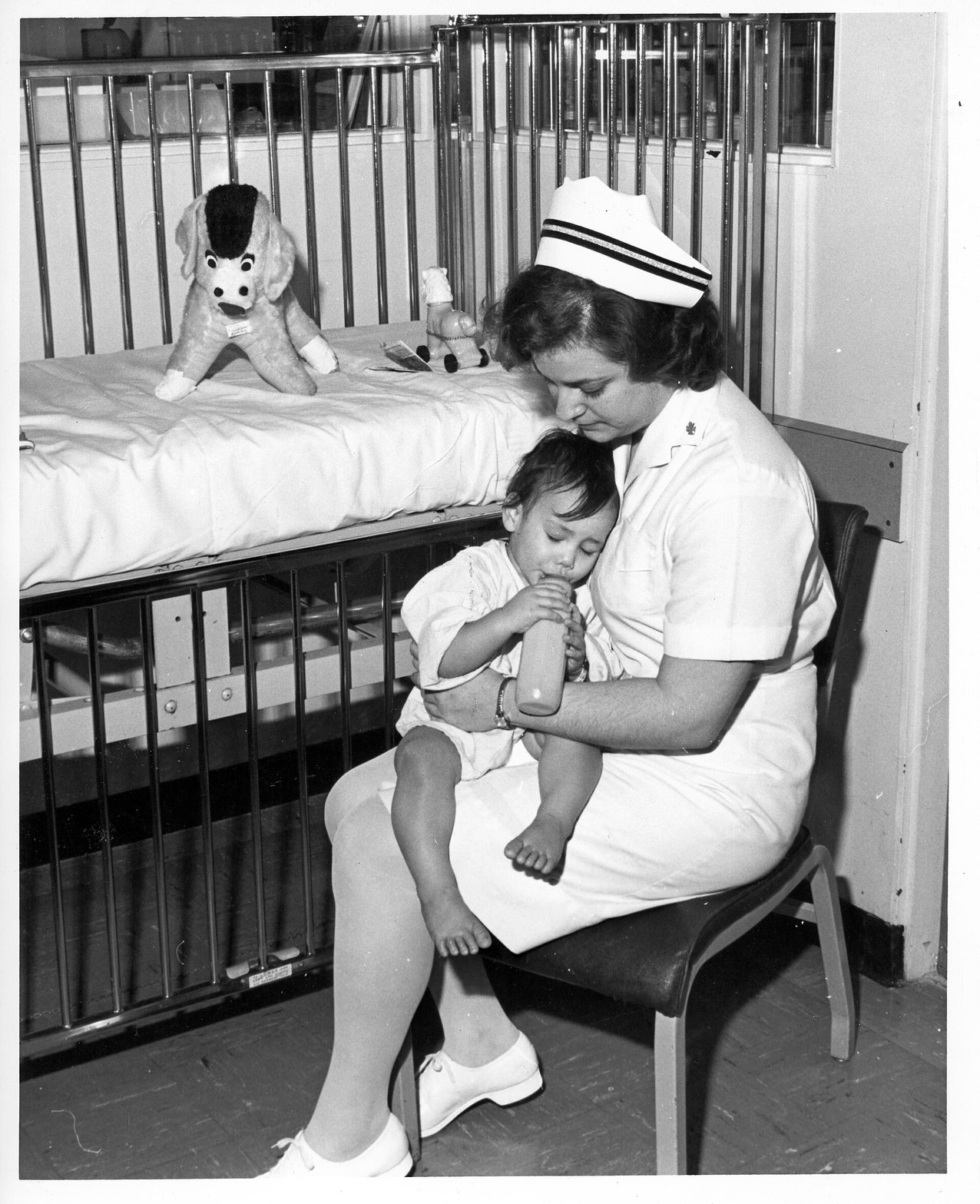 Fotografía en blanco y negro de una enfermera con uniforme y cofia blancos sentada junto a una cuna de hospital, con un niño pequeño en brazos que bebe de un biberón