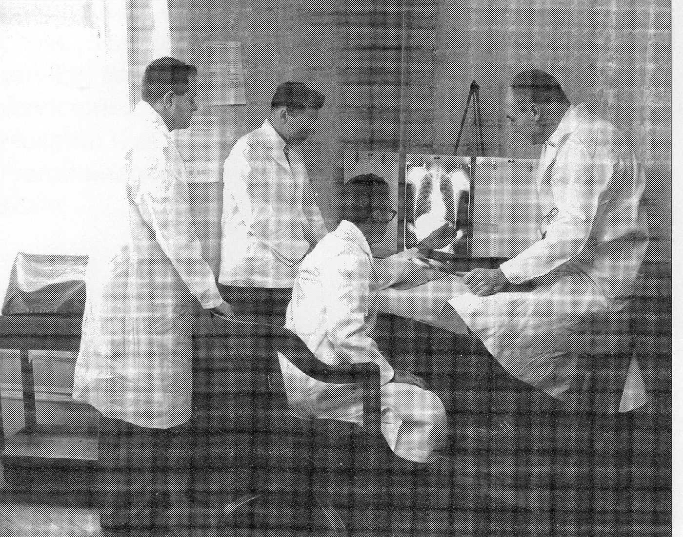 Fotografía en blanco y negro de cuatro médicos con bata blanca examinando una radiografía de tórax junto a un paciente sentado
