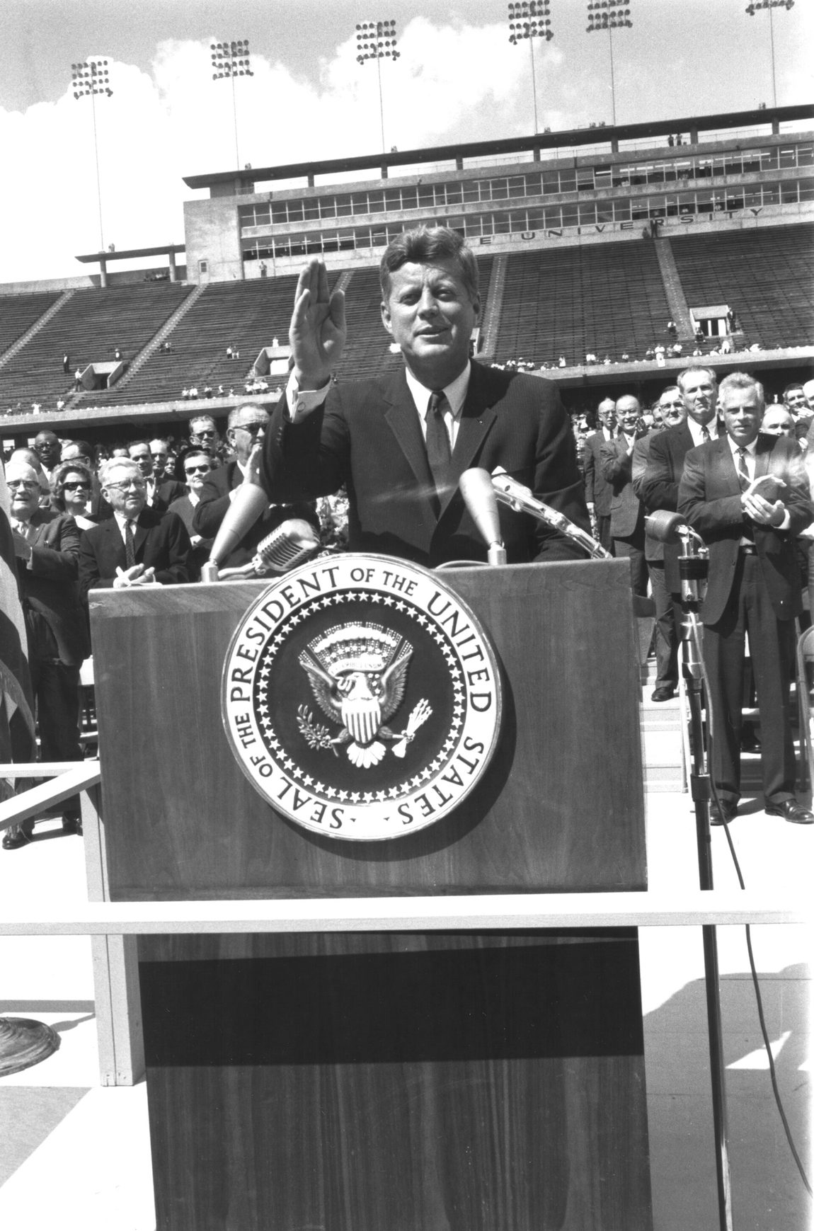 Fotografía en blanco y negro de John F. Kennedy hablando en un atril con el sello presidencial ante un estadio lleno