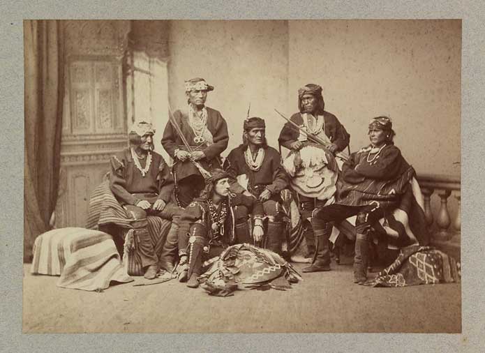 Fotografía antigua en tono sepia de un grupo de hombres indígenas con ropas tradicionales, sentados y de pie en un estudio fotográfico, junto a un hombre blanco