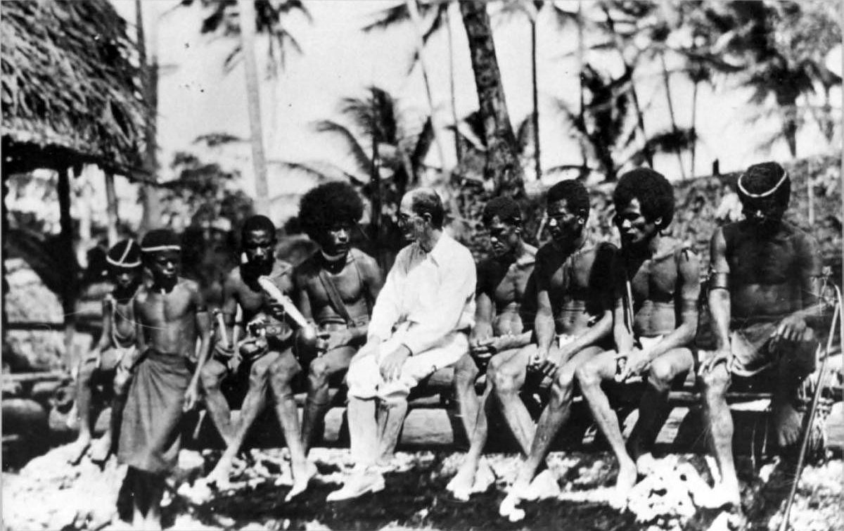 Fotografía en blanco y negro de un hombre vestido de blanco sentado en un banco junto a un grupo de hombres trobriandeses, delante de una casa con techo de hojas y palmeras