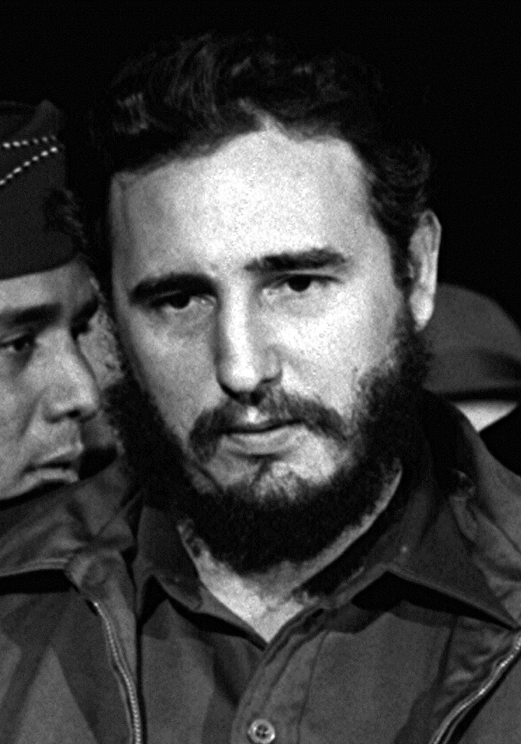 Fotografía en blanco y negro de un hombre con barba y uniforme militar, junto a otras personas en primer plano, en un aeropuerto