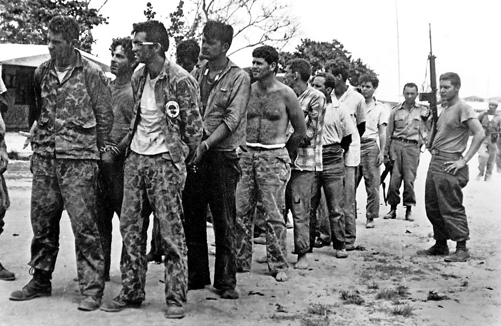 Fotografía en blanco y negro de un grupo de hombres con ropa militar, algunos con las manos en la cabeza, en fila y vigilados por soldados en un terreno arenoso