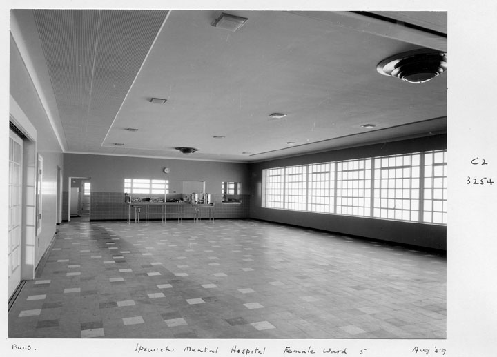 Fotografía en blanco y negro de una sala amplia y vacía de un hospital psiquiátrico, con suelo de baldosas y ventanales