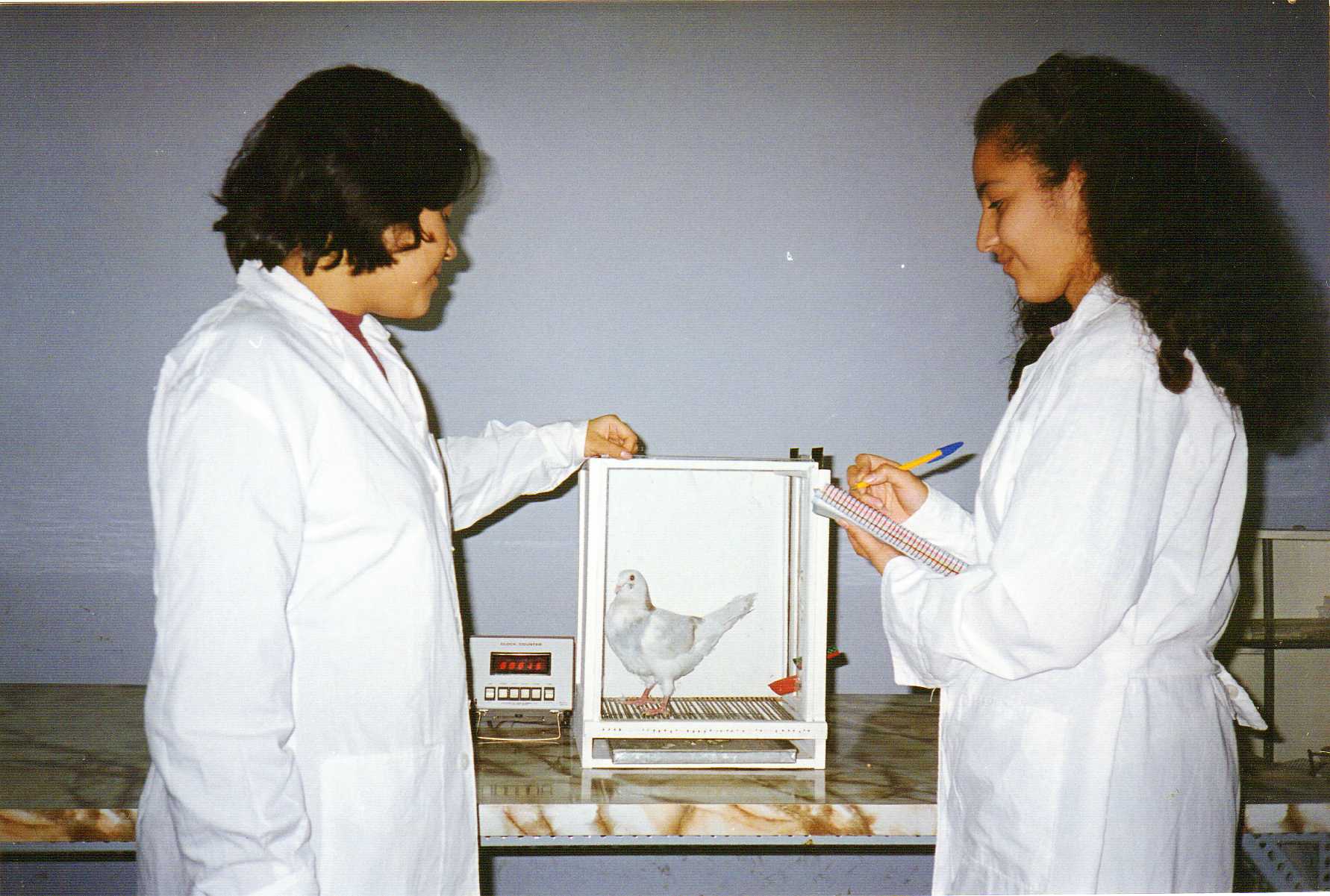 Fotografía en color de dos estudiantes con bata blanca en un laboratorio, una de ellas toma notas junto a una caja de metacrilato con una paloma blanca dentro