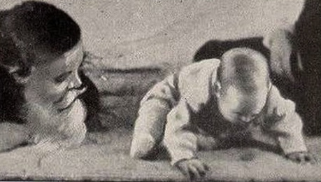 Fotograma en blanco y negro de un bebé gateando sobre una colchoneta junto a un hombre con una máscara de Papá Noel