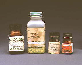 Fotografía en color de cuatro frascos de farmacia de distintos tamaños con etiquetas de placebo, uno de ellos lleno de pastillas amarillas