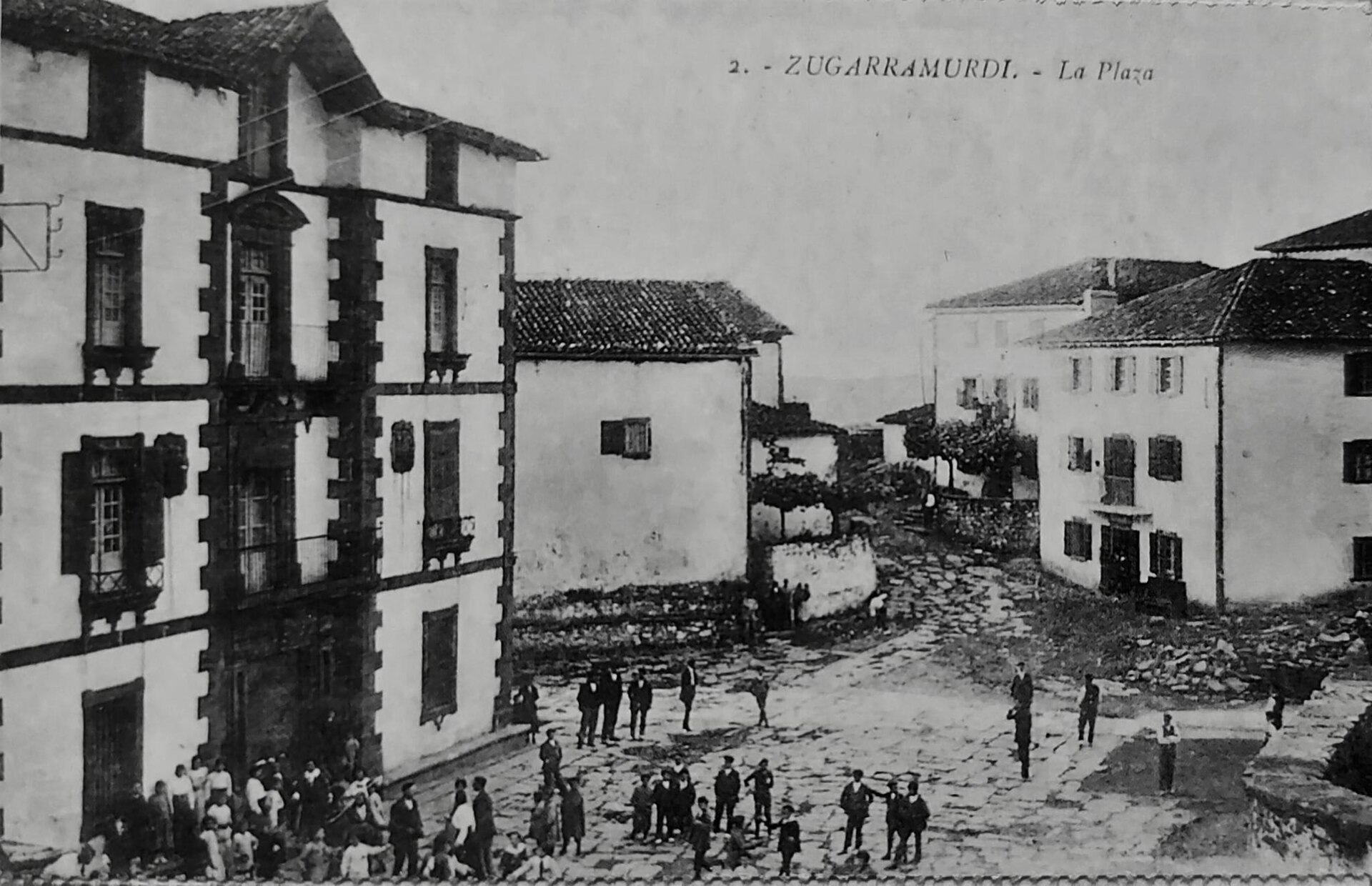 Postal antigua en blanco y negro de la plaza de un pueblo con casas de piedra de varias plantas y un grupo de vecinos en la calle