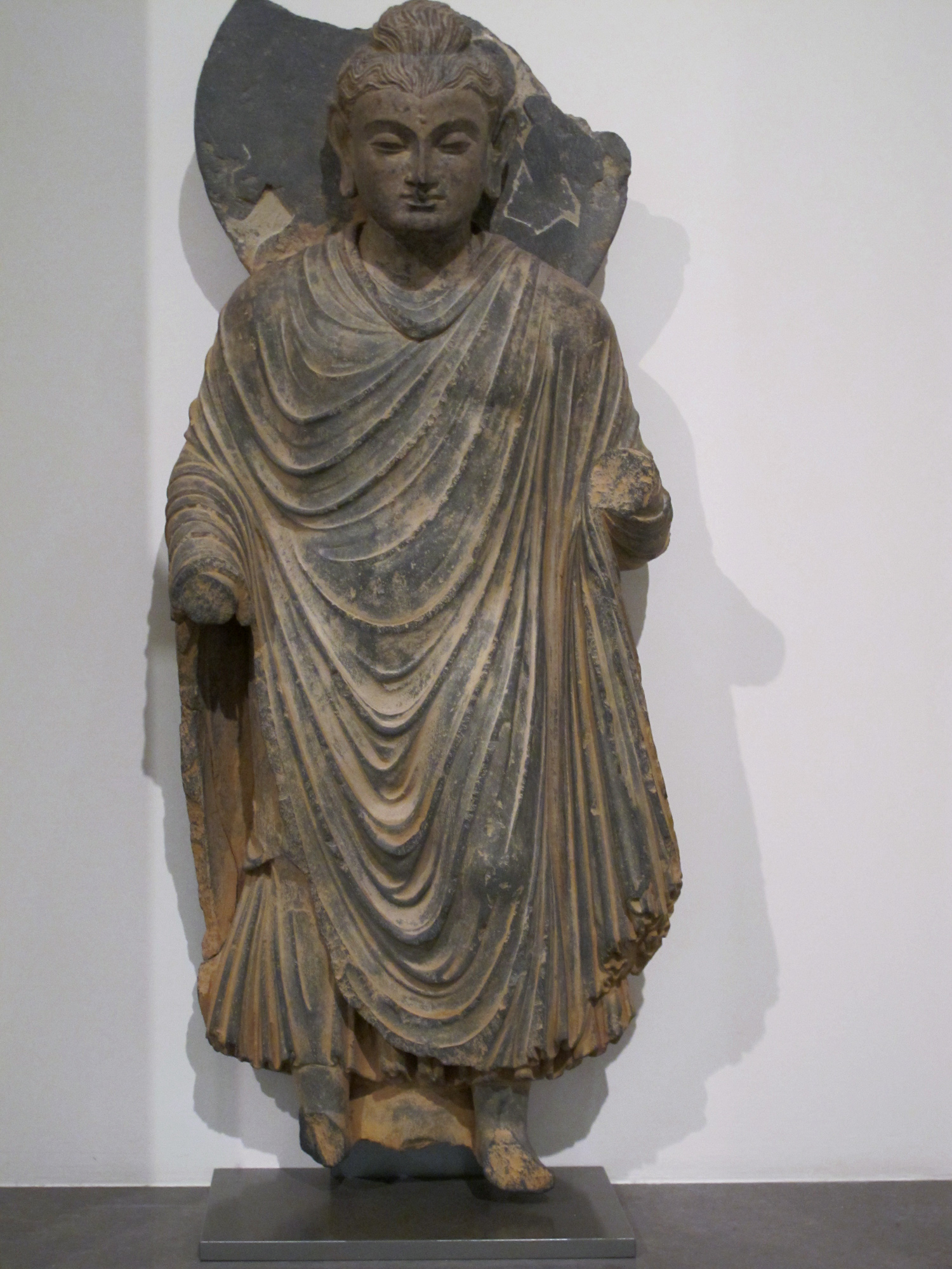 Estatua de piedra gris de un hombre de pie con el pelo recogido en un moño, vestido con un manto de pliegues profundos que le cubre ambos hombros