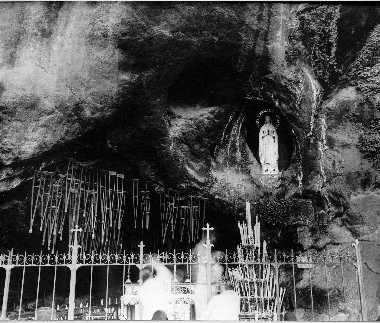 Fotografía antigua de la gruta iluminada por velas, con la estatua de la Virgen en una hornacina y decenas de muletas colgadas de un cable sobre la entrada